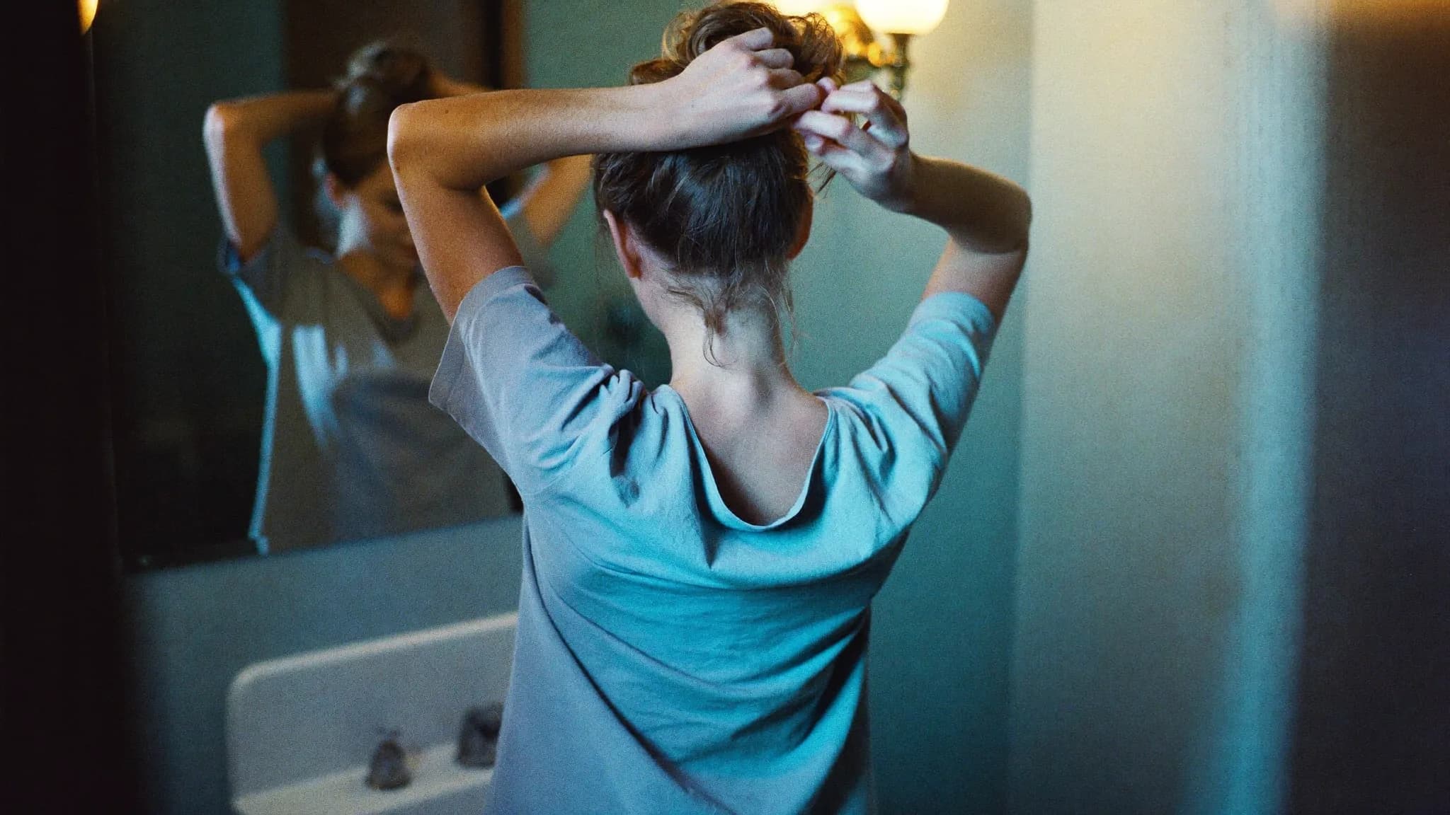 A woman gathering her hair up in a bathroom mirror, warm lamp light behind her